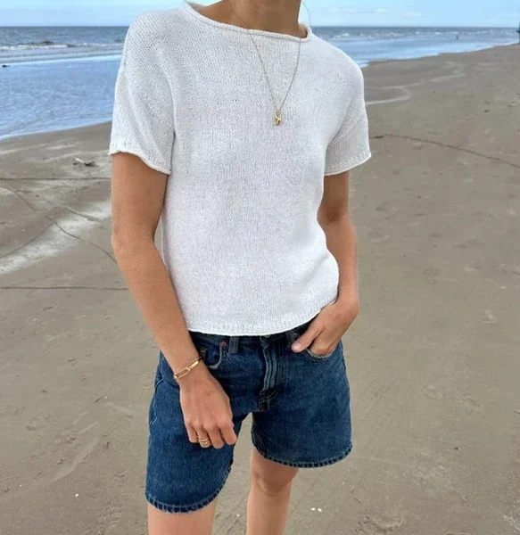 En person står på en sandstrand iført en kortermet hvit skjorte, denimshorts, et halskjede og et armbånd, med havet og himmelen i bakgrunnen.