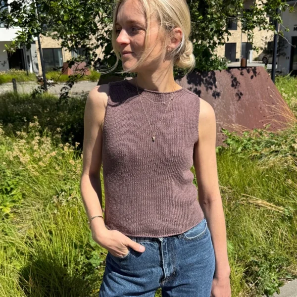 En kvinne står utendørs i sollys, iført den ermeløse, brune Ivy-toppen og blå jeans, med grøntområder og bygninger i bakgrunnen.