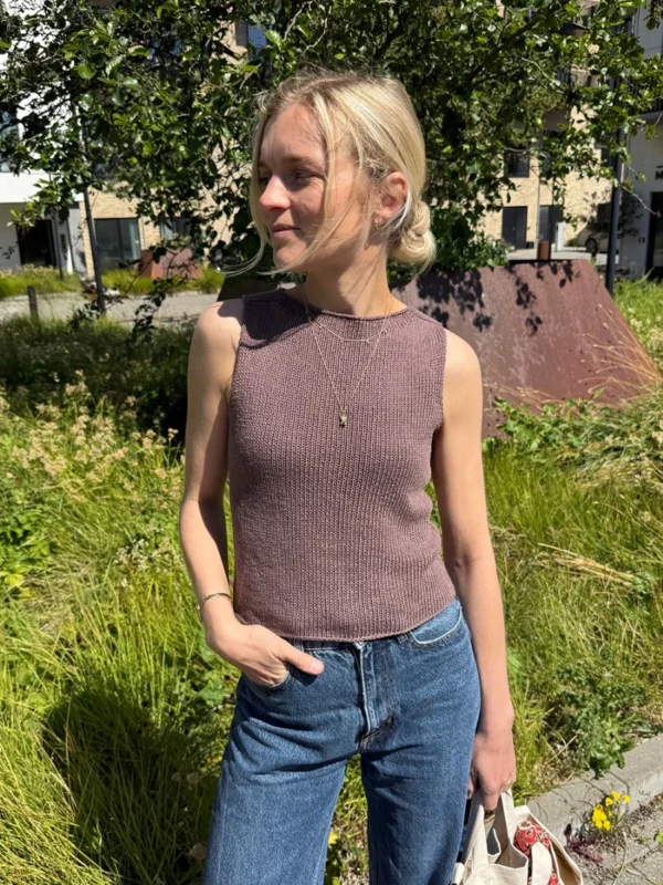 En kvinne står utendørs i sollys, iført den ermeløse, brune Ivy-toppen og blå jeans, med grøntområder og bygninger i bakgrunnen.