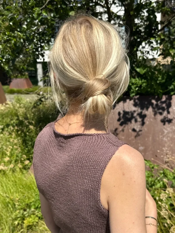 En person med lysblondt hår i en lav knute står utendørs blant grønne planter og sollys, iført den ermeløse, strikkede Ivy Top.