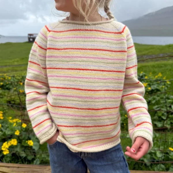 Barn står utendørs iført kremfarget genser med fargerike striper og blå jeans, med grønne planter og gule blomster i bakgrunnen.