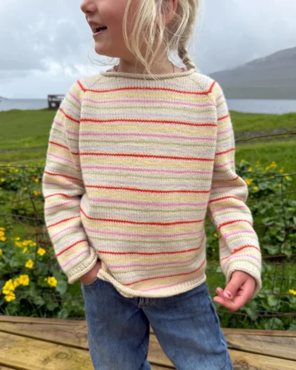 Barn står utendørs iført kremfarget genser med fargerike striper og blå jeans, med grønne planter og gule blomster i bakgrunnen.