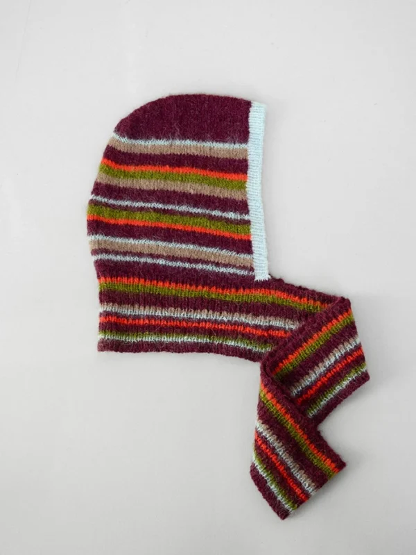 2507_11 Snuggle hood er en stripete strikklue med rødbrune, grønne, grå, røde og gule striper og en lyseblå kant, vist liggende på en hvit bakgrunn.