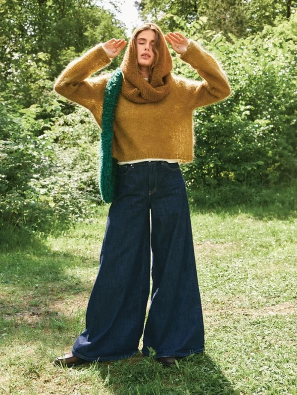 Utendørs på gresset har en person på seg genser 2507_03 Rookie med hette, blå jeans med vide ben, brune sko og et grønt skjerf over den ene skulderen. I bakgrunnen skimtes trær og busker.