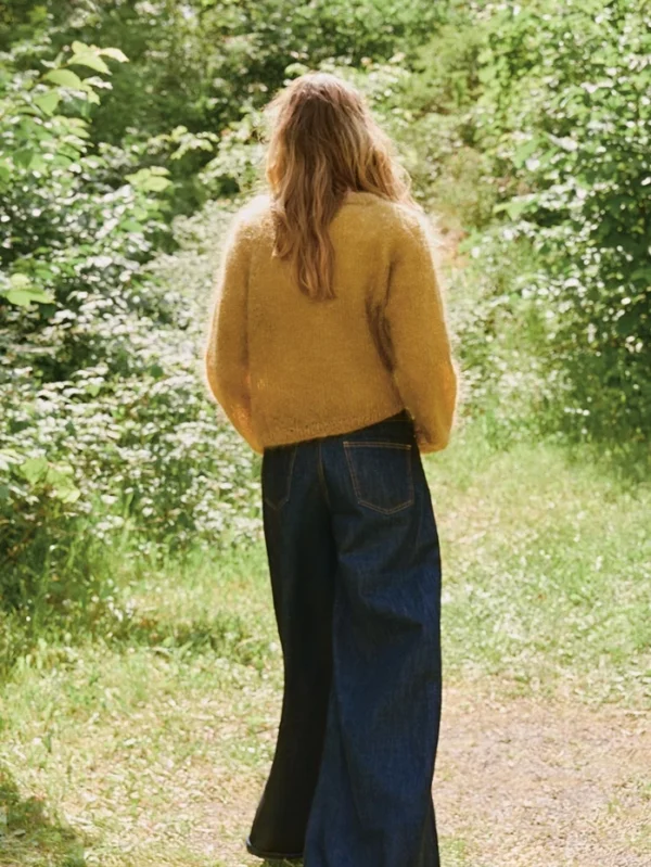 En person med langt hår, iført Rookie-genseren 2507_03 Rookie i gult og jeans med vide ben, står på en gressbelagt sti omgitt av grønne planter, vendt bort fra kameraet.