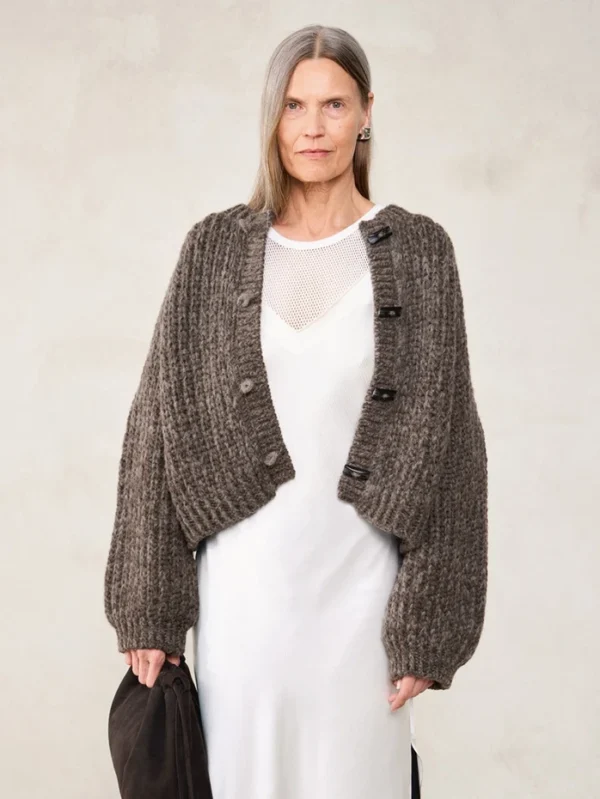 En kvinne med langt, grått hår har på seg den brune strikkekofta 2507_04 Chunky jacket style over en hvit kjole, mens hun står mot en lys bakgrunn og holder en mørk veske.