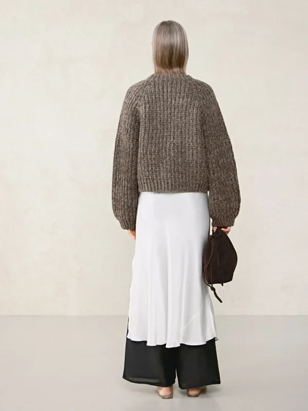 En person med rett gråblondt hår, sett bakfra, har på seg den brune genseren 2507_04 Chunky jacket-stil over en lang hvit kjole med svart nederkant og bærer en mørk veske.