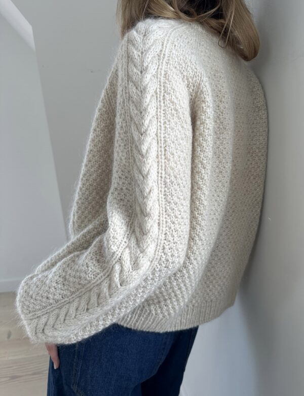 En person står med ryggen til kameraet ved en hvit vegg, iført Siri Cardigan i chunky offwhite og mørkeblå jeans.