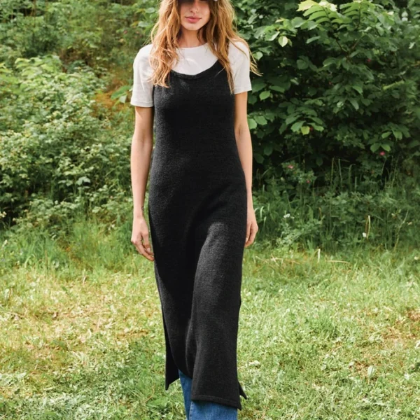 En kvinne iført 2507_02 Emerson-kjole over en hvit t-skjorte, blå jeans og en NY-baseballcaps står på gresset med grønne trær i bakgrunnen.