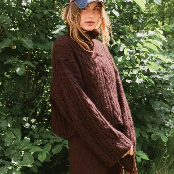 En person står utendørs foran grønt løvverk, iført en løs, brun 2507_01 Brady-genser, et matchende skjørt og en fargerik baseballcap.