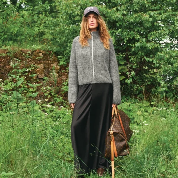 En kvinne står i høyt gress iført 2507_10 Tia-jakke med glidelås, svart skjørt, hette og brune støvler, og holder en stor brun veske med lærrem. Bakgrunnen er dekket av grønt løvverk.