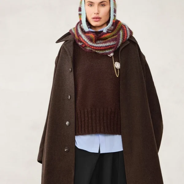 Kvinne iført stor brun frakk, brun genser, lyseblå skjorte, svarte bukser og 2507_11 Snuggle hood flerfarget strikket skjerf og hodeplagg mot en nøytral bakgrunn.