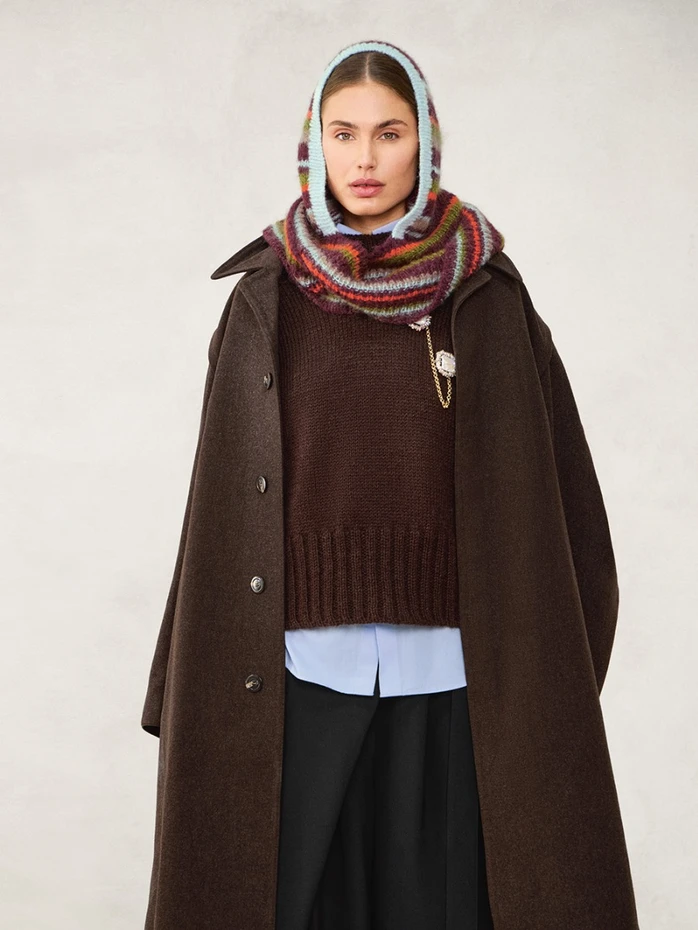 Kvinne iført stor brun frakk, brun genser, lyseblå skjorte, svarte bukser og 2507_11 Snuggle hood flerfarget strikket skjerf og hodeplagg mot en nøytral bakgrunn.