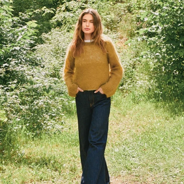En kvinne står utendørs på en gressbelagt sti, omgitt av grøntområder og sollys, iført genser 2507_03 Rookie og mørkeblå jeans.