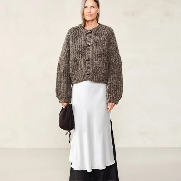 En kvinne står foran en ensfarget bakgrunn iført 2507_04 Chunky-jakken over en lang, hvit kjole med svarte detaljer, og holder en liten, brun veske.