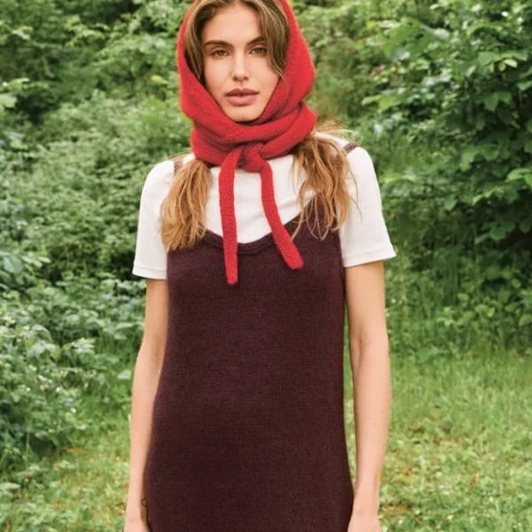 En kvinne står utendørs med grønne planter bak seg, iført 2507_07 Tie the hood - et rødt skjerf utformet som en hette - over en hvit t-skjorte og en mørk, ermeløs kjole.