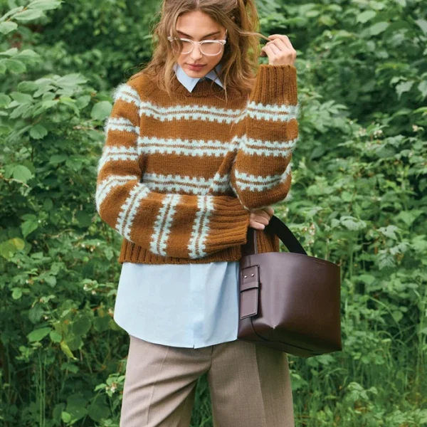 En person iført den brunstripete Ridge-genseren 2507_08 over en blå skjorte og beige knelange shorts står utendørs med en mørkebrun håndveske i hånden, omgitt av grønt løvverk.