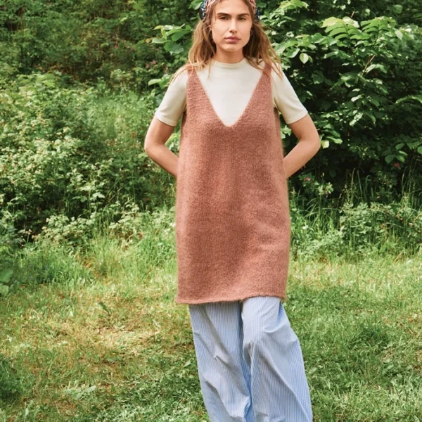 En kvinne står på gresset i beige T-skjorte, kjolen 2507_09 Meadow (brun, uten ermer), blåstripete bukser med vide ben, brune sko og et rosa mønstret skjerf med grønt løvverk i bakgrunnen.