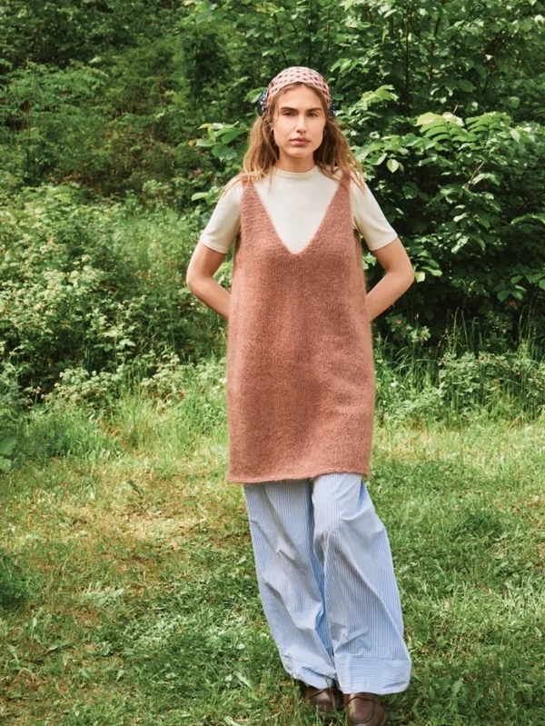 En kvinne står på gresset i beige T-skjorte, kjolen 2507_09 Meadow (brun, uten ermer), blåstripete bukser med vide ben, brune sko og et rosa mønstret skjerf med grønt løvverk i bakgrunnen.