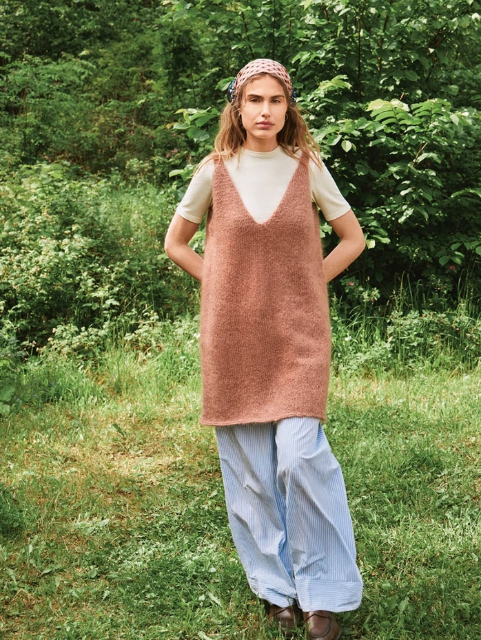 En kvinne står på gresset i beige T-skjorte, kjolen 2507_09 Meadow (brun, uten ermer), blåstripete bukser med vide ben, brune sko og et rosa mønstret skjerf med grønt løvverk i bakgrunnen.