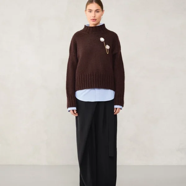 En kvinne med bakoverbøyd hår og nøytralt ansiktsuttrykk står opp mot en vanlig vegg iført 2507_05 Mindy-genser i mørkebrunt over en lyseblå skjorte, vide, svarte bukser og svarte sko.