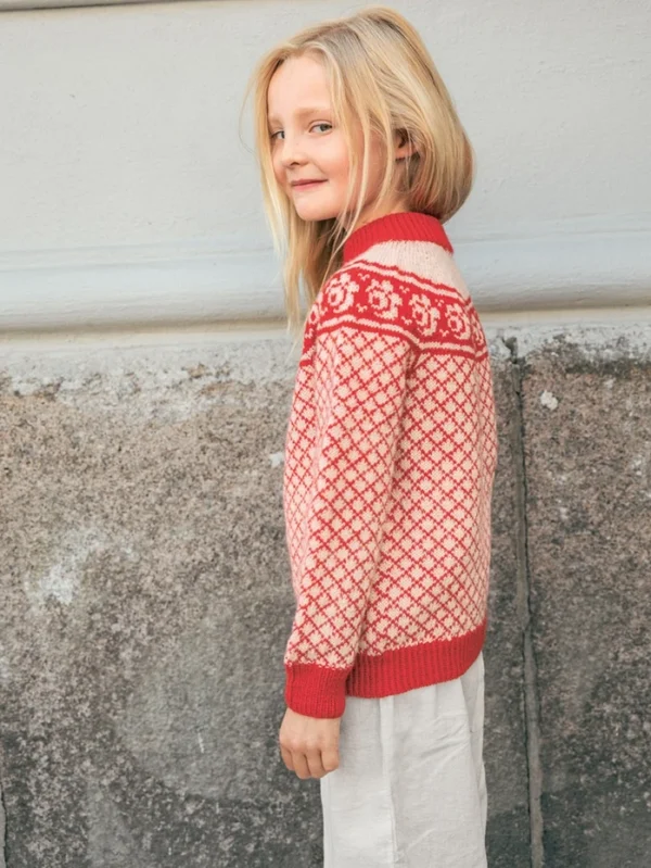 En ung jente med langt, blondt hår står sidelengs mot en vegg av strukturert stein og gips, iført 2509_01 Blome junior-genser og lyse bukser.