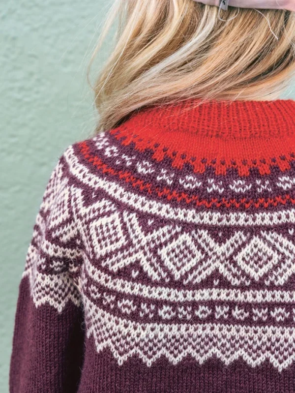 En person med blondt hår har på seg Marius junior-genseren 2509_02, som har et rødt, hvitt og lilla fair isle-mønster, mens hun står foran en vegg med grønn tekstur.