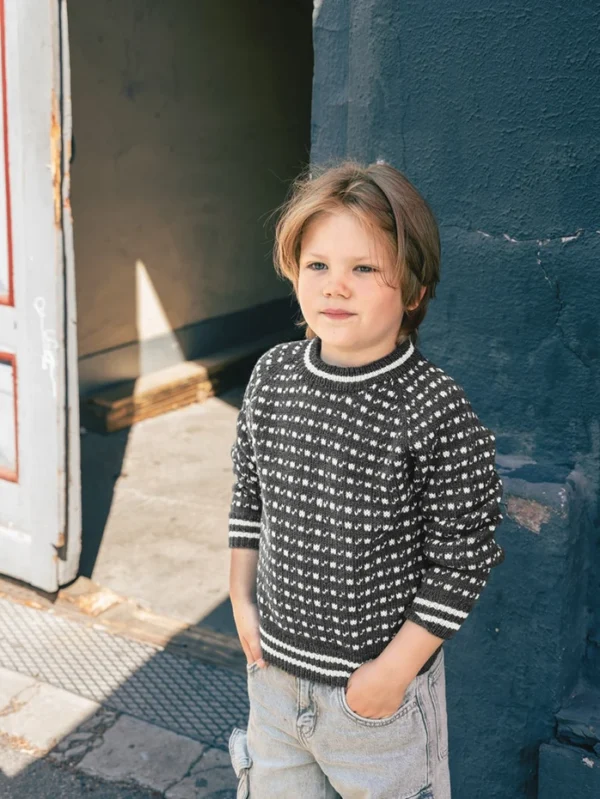 Et lite barn med lysebrunt hår står ute foran en mørk vegg, iført 2509_03 Islender junior-genser med kraftig strikkemønster og lyse bukser, med hendene i lommene.
