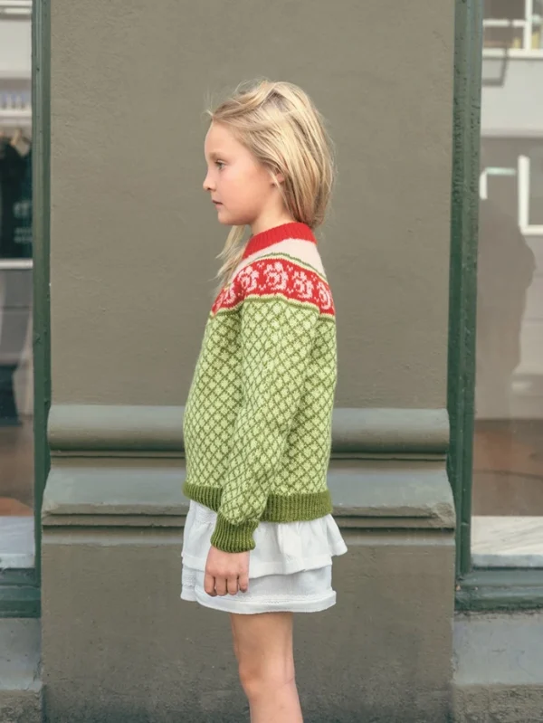 En ung jente står i profil mot en grå vegg, iført en 2509_05 Blome junior grønnmønstret genser med rødt og hvitt bærestykke over et lyst skjørt.