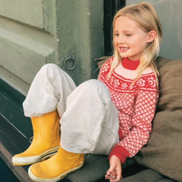 Et lite barn med langt blondt hår sitter i vinduskarmen, smiler og ser til siden, kledd i 2509_01 Blome junior-genser, løse hvite bukser og gule støvler.
