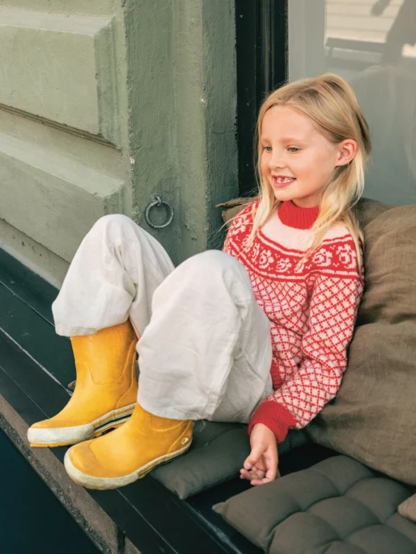 Et lite barn med langt blondt hår sitter i vinduskarmen, smiler og ser til siden, kledd i 2509_01 Blome junior-genser, løse hvite bukser og gule støvler.
