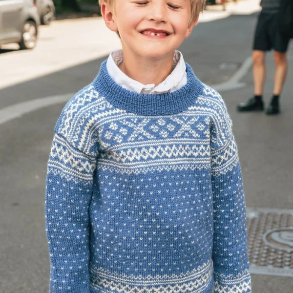 En ung, blond gutt smiler med lukkede øyne, iført jeans og 2509_04 Setesdal juniors blå og hvite genser, stående i en bygate i dagslys.