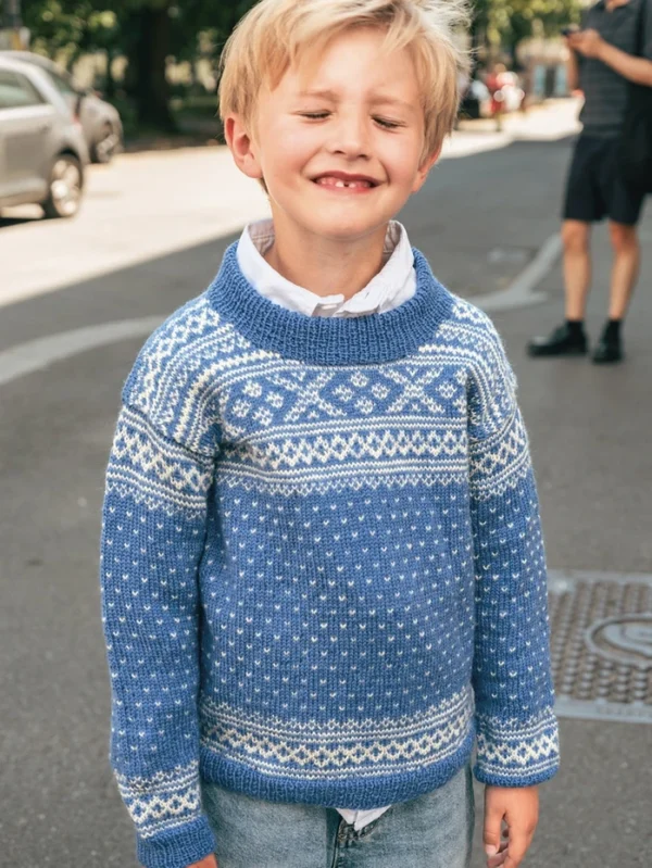 En ung, blond gutt smiler med lukkede øyne, iført jeans og 2509_04 Setesdal juniors blå og hvite genser, stående i en bygate i dagslys.