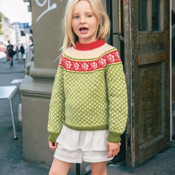En ung jente står utenfor en bygning, iført 2509_05 Blome junior grønnmønstret genser med røde og hvite detaljer, sammen med hvite shorts og gule støvler.
