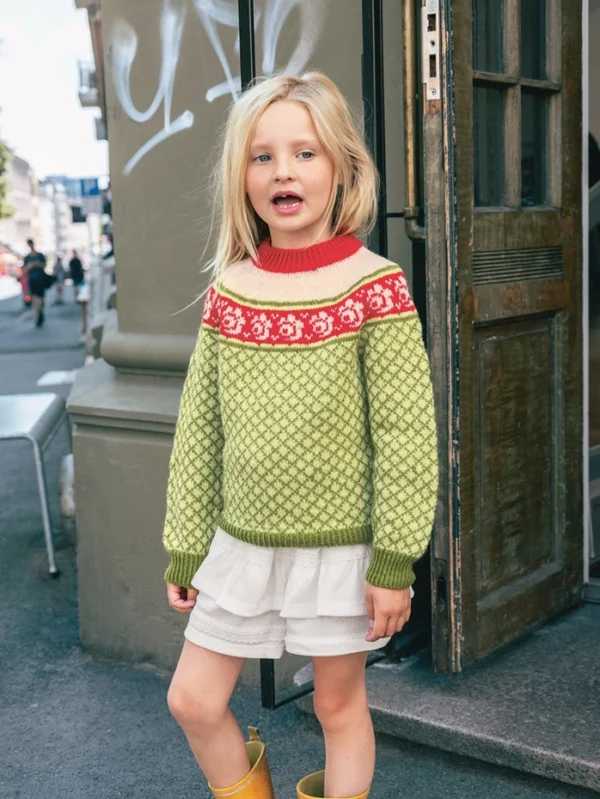 En ung jente står utenfor en bygning, iført 2509_05 Blome junior grønnmønstret genser med røde og hvite detaljer, sammen med hvite shorts og gule støvler.