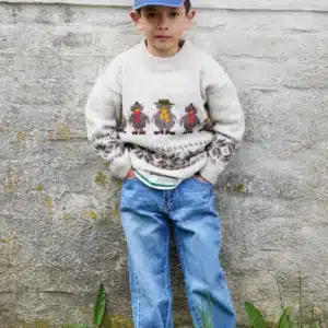 En ung gutt i grå genser med fuglemønster, blå jeans og blå caps står med hendene i lommene mot en strukturert, lys vegg.